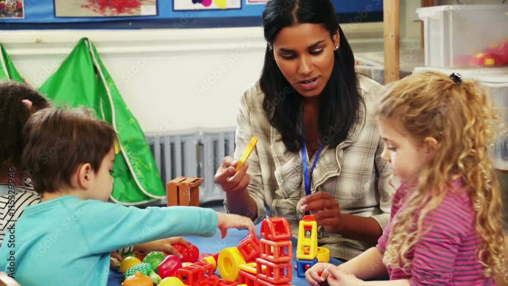 Teacher playing with building blocks and other toys at a classroom table with her nursery students.