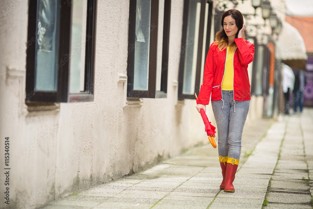 Smiling young woman is walking in a city on a rainy day. She is wearing red raincoat, rubber boots and holding a red umbrella.