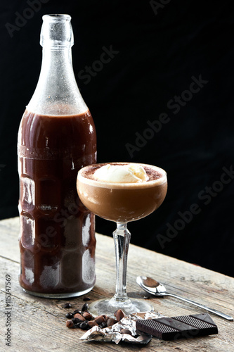 Alcoholic cocktail Ice Scream Russian is served in a glass bottle and goblet on wooden table on black background