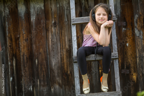 Little naughty girl with headphones sitting on a wooden stepladder in the village outdoors. photo