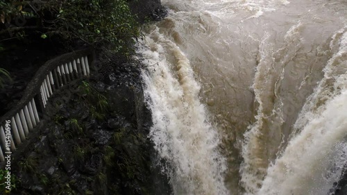 A high-angle view of turbulent muddy water rushing over dark wet rocks at the Pailon del Diablo near Banos, Ecuador, white foam and rapids, a stone railing and lush green foliage on the left bank.