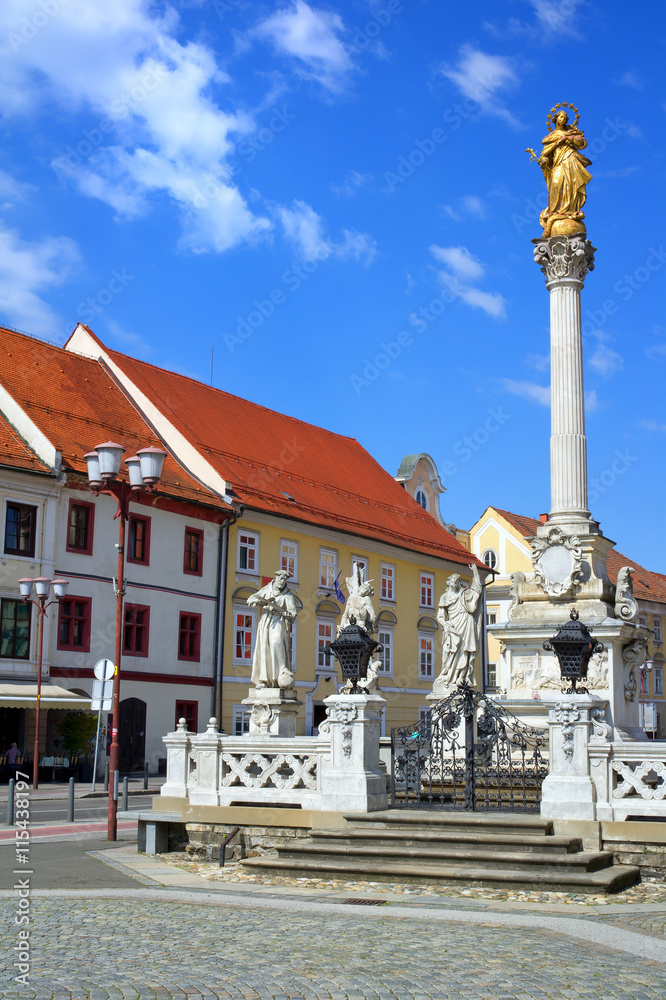 Fototapeta premium Plague column (Kuzno znamenje) at Main Square (Glavni Trg) of the city of Maribor in Slovenia, Europe. Historical center with religious sculpture. Street in the city and town. Bright sunny weather.