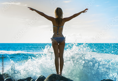 Young beautiful woman standing on the stones nearly waves splash with sea and sky background. Back view photo