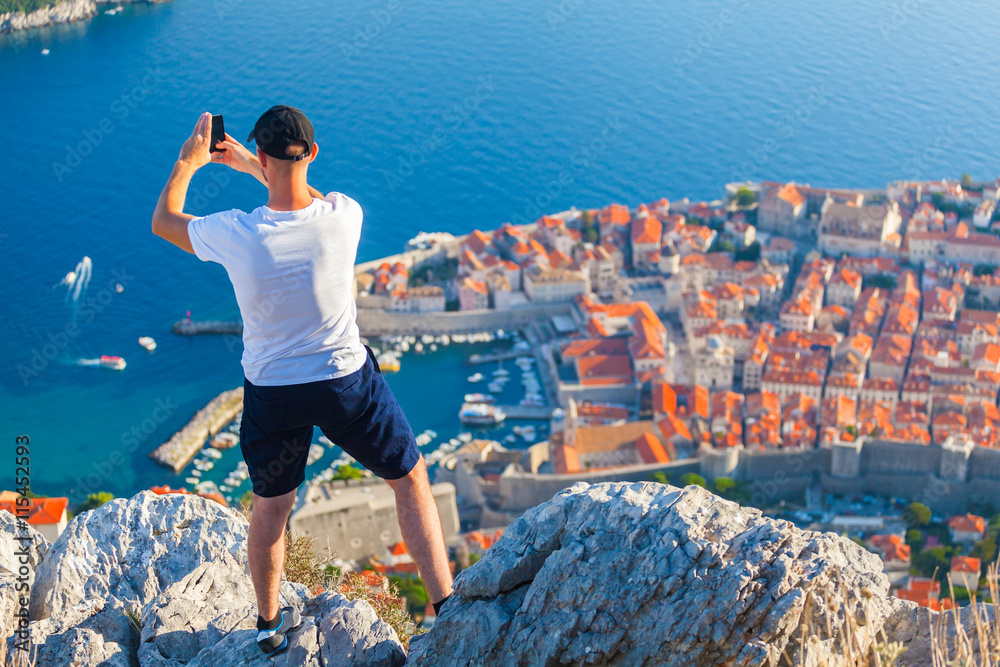 Male traveller make a panoramic photo of the old city of Dubrovnik from the mountain to his phone for Instagram or other social networks. Travel to Croatia. Summer vacation.