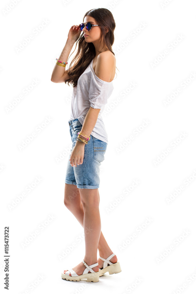 Woman stands and touches sunglasses. White tunic and denim shorts. Plain clothes and footwear. When sun shines brightly.