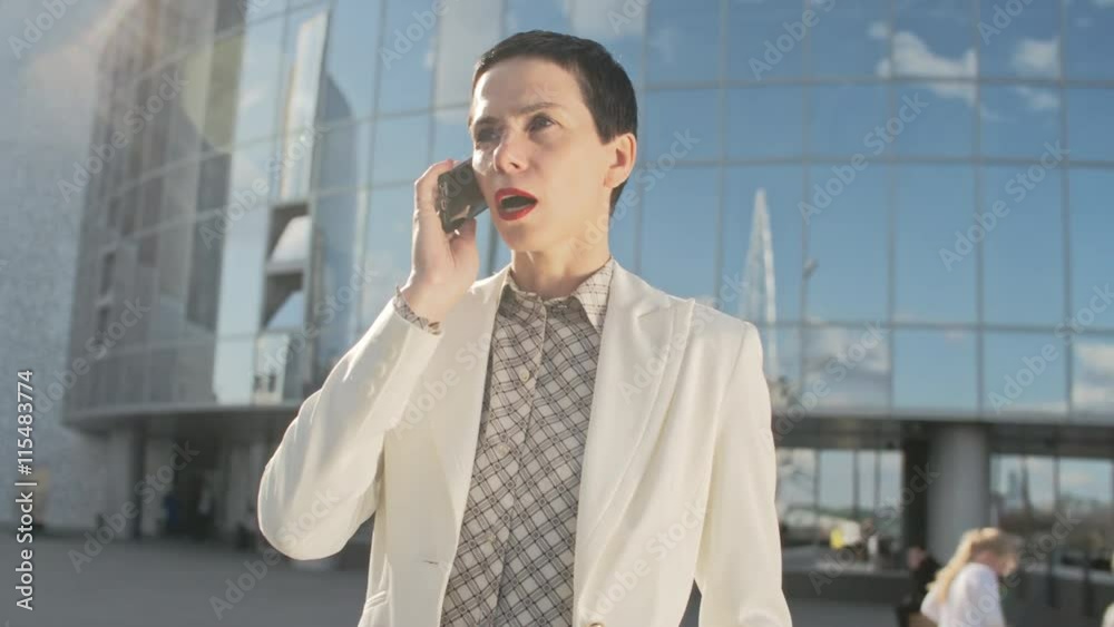 Mid-adult businesswoman with short dark hair speaking on the phone while standing near modern glass office center Mid-adult businesswoman with short dark hair speaking on the phone while standing near modern glass office center