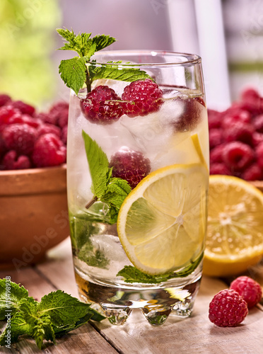 On wooden boards is cocktail glass with berries lemon and ice cubes. Alcohol country cocktail number eighty two with berry bowl mint and ice cubes.