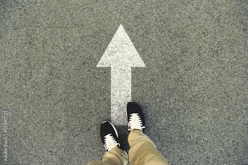 Forward arrow painted on an asphalt road. Top view of the legs and shoes. POV