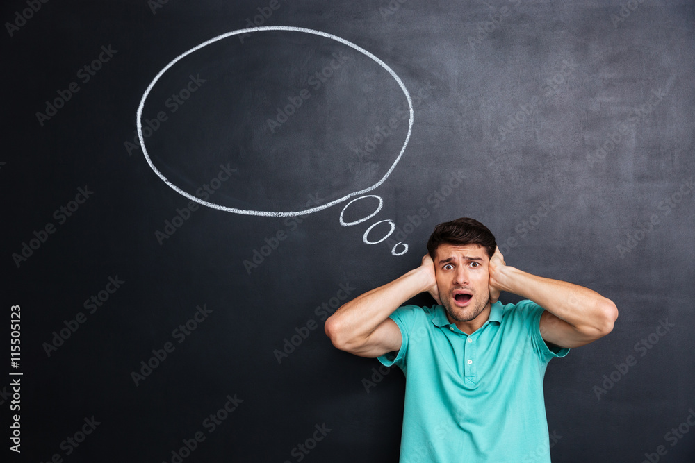 Shocked stressed man covered ears by hands over chalkboard background