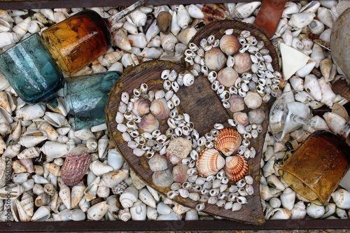 Composition of heart made of wood and shells surrounded by broken colorful glasses. At the background many shells.