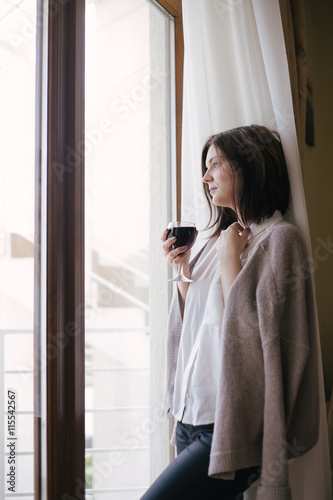 Young beautiful woman in a sweater drinking red wine near big window in a cozy room