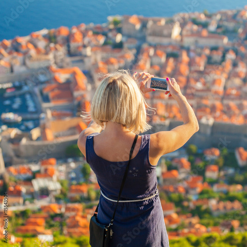 Female traveller make a panoramic photo of the old city of Dubrovnik from the mountain to her phone for Instagram or other social networks. Travel to Croatia. Summer vacation.