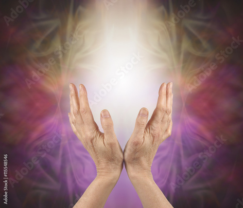 Pranic healer sensing energy  -   female hands reaching up into a soft white light with a gold pink and purple energy formation behind and copy space 