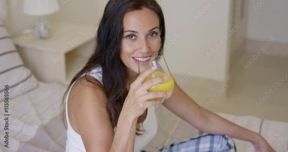 Smiling healthy young woman drinking a glass of freshly squeezed orange juice as she sits on her bed in the morning Smiling healthy young woman drinking a glass of freshly squeezed orange juice as she sits on her bed in the morning