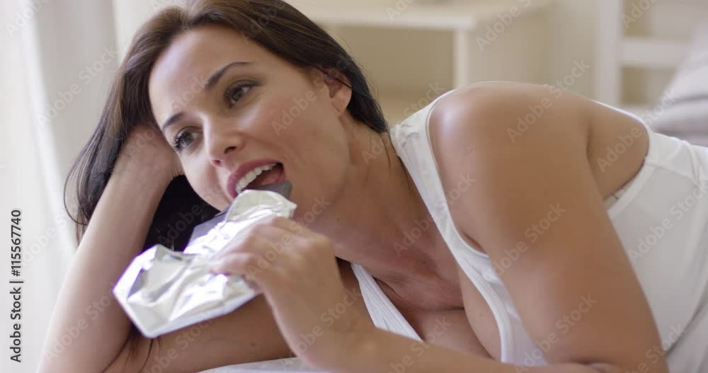 Adorable middle aged woman eating bar of dark chocolate while laying in her pijama on bed in the morning Adorable middle aged woman eating bar of dark chocolate while laying in her pijama on bed in the morning
