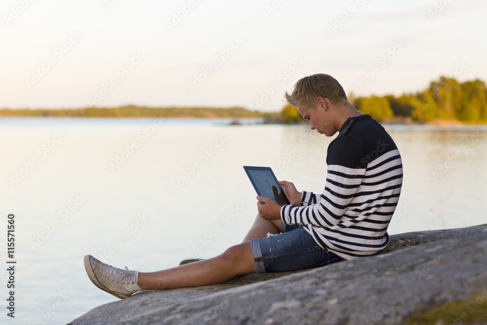 Sweden, Ostergotland, Teenage boy (16-17) sitting on rock and using digital tablet Sweden, Ostergotland, Teenage boy (16-17) sitting on rock and using digital tablet
