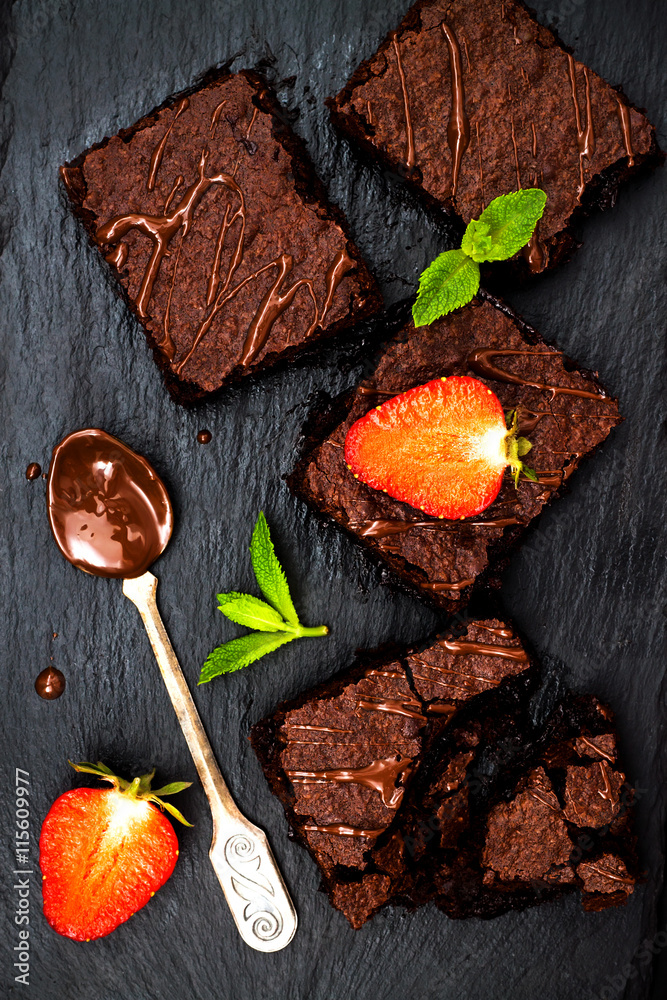 Homemade dark chocolate brownies decorated with strawberries and mint leaves over black slate background, top view