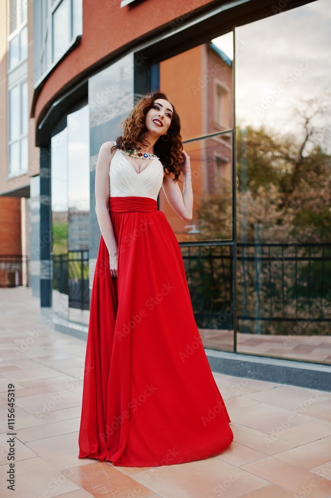 Portrait of fashionable girl at red evening dress posed background mirror window of modern building