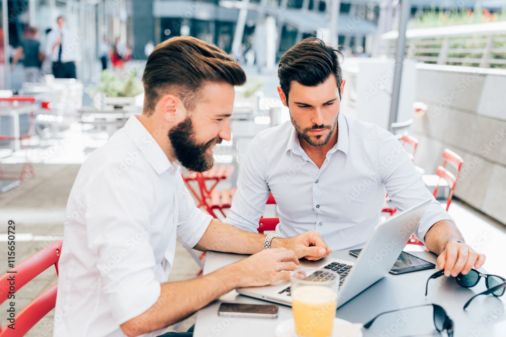 Two young contemporary businessmen working outdoor sitting in a bar using a smart phone and a notebook, both smiling - technology, business, work concept - copy space on left
