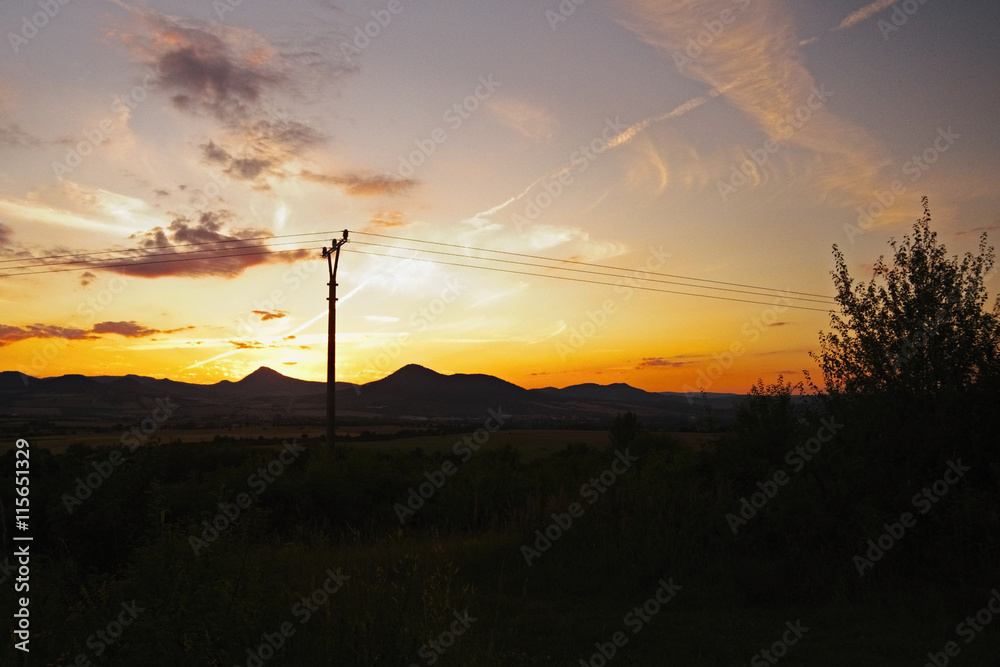 Fototapeta premium romantic sunset in the region Ceske stredohori in czech landscape with fields, pylons and mountains Milesovka and Lovos
