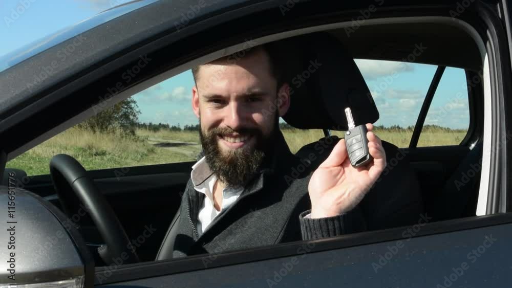 young handsome hipster man sits in the car and holds key from the car and smiles to camera - countryside - sunny weather 