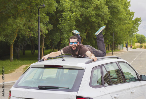 Young man with beard and sunglasses lying and shouting on the roof of a car.