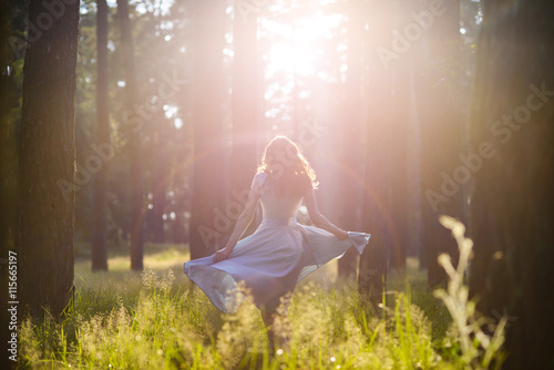 Obraz na plátně Beautiful young woman wearing elegant light blue dress standing in the forest with rays of sunlight beaming through the leaves of the trees