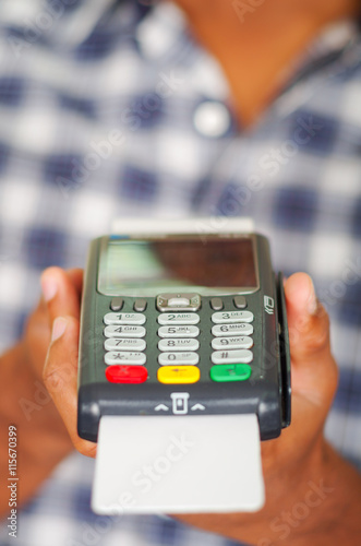 Man wearing blue white square pattern shirt holding up credit card payment terminal in front of camera, closeup angle