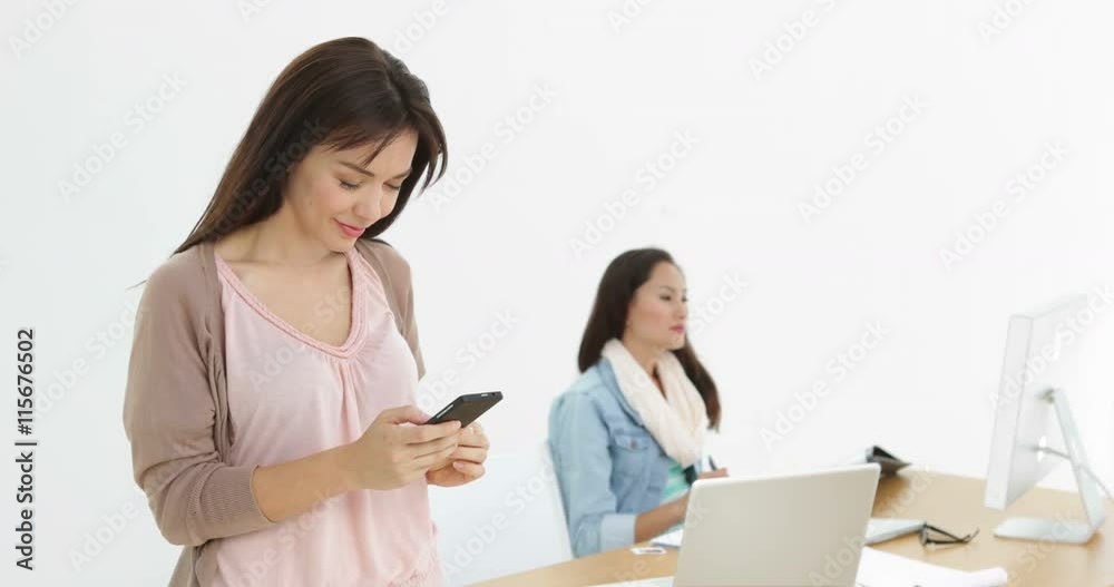 Creative businesswoman sending a text with colleague behind her Creative businesswoman sending a text with colleague behind her