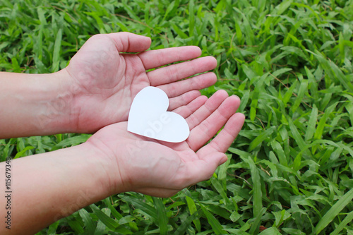 Two hands holding white heart with green grass background.