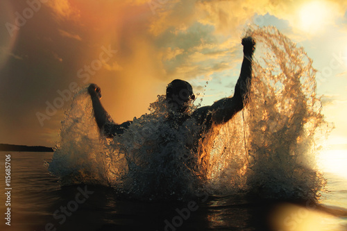 Fotografia Strong and athletic man jumps out of the water at sunset, flying a lot of splash