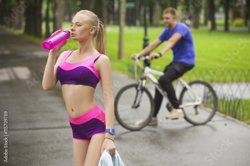 Beautiful girl drinks water and the other hand holding the towel . In the background the guy on the bike