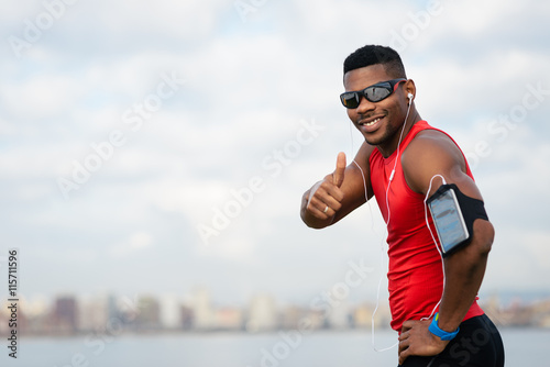 Fototapeta Naklejka Na Ścianę i Meble -  Positive urban black athlete celebrating running workout success towards city skyline background. Cheerful man doing successful thumbs up gesture.