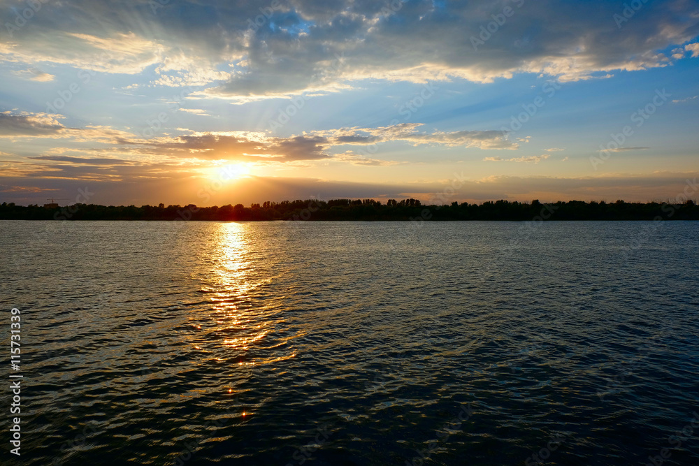 Orange setting sun over water surface with ripples and tree tops on far coast Orange setting sun over water surface with ripples and tree tops on far coast