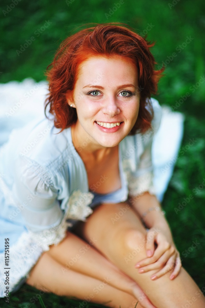 red-haired young woman in elegant blue dress sitting on the grass and smiling, looking into the camera red-haired young woman in elegant blue dress sitting on the grass and smiling, looking into the camera
