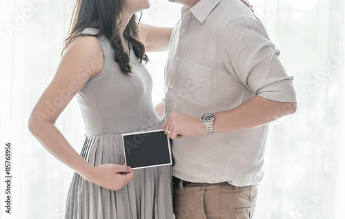 Pregnant woman and husband holding ultrasound photo of baby in front of the window with high key light