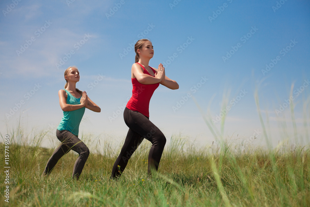 Two women doing yoga outdoors on blue sky background posing on g Two women doing yoga outdoors on blue sky background posing on g