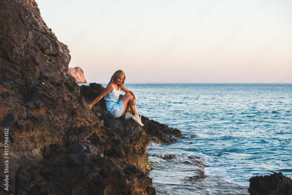Young blond woman tourist in blue dress sittig on rocks and looking at the sea. Alanya, Mediterranean region, Turkey.
