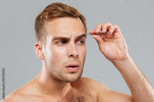 Closeup of concentrated young man removing eyebrow hairs with tweezers