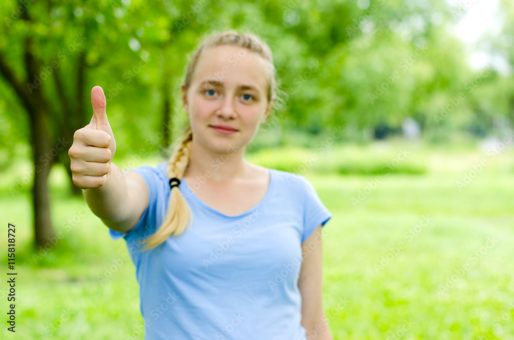 Young blond girl in the blue shirt shows positive sign in the park. She is pretty and active. Sunny positive mood and emotions.
