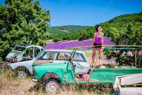 Tableau sur toile Femme en short dans une casse de vieilles voitures devant les champs de lavandes