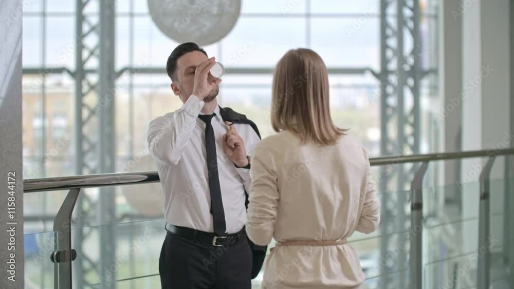 Businessman standing in the corridor of business center, holding jacket on his shoulder, drinking coffee and talking with female colleague Businessman standing in the corridor of business center, holding jacket on his shoulder, drinking coffee and talking with female colleague