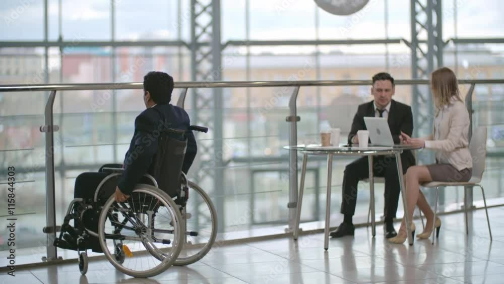 Disabled businessman sitting on wheelchair and talking on the phone; man and woman sitting at the glass table with laptop and talking Disabled businessman sitting on wheelchair and talking on the phone; man and woman sitting at the glass table with laptop and talking