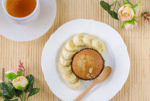 Flat lay of piece of fresh delicious banana cup cake with sliced fresh banana on white ceramic plate over bamboo placemat with black tea in white ceramic cup decorated with light pink small roses.