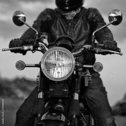 Black and white image of the front of a motorbike showing headlamp and mirrors. Which adult male in leathers.