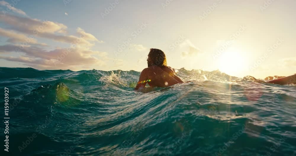 Surfer paddling over blue ocean wave at sunset in slow motion