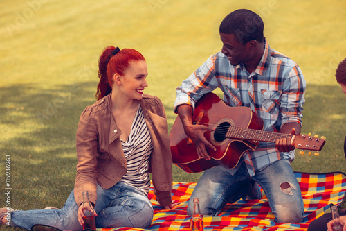 Happy couple resting on picnic in parklane. Afro-American man playing guitar and singing songs to his girl-friend. photo