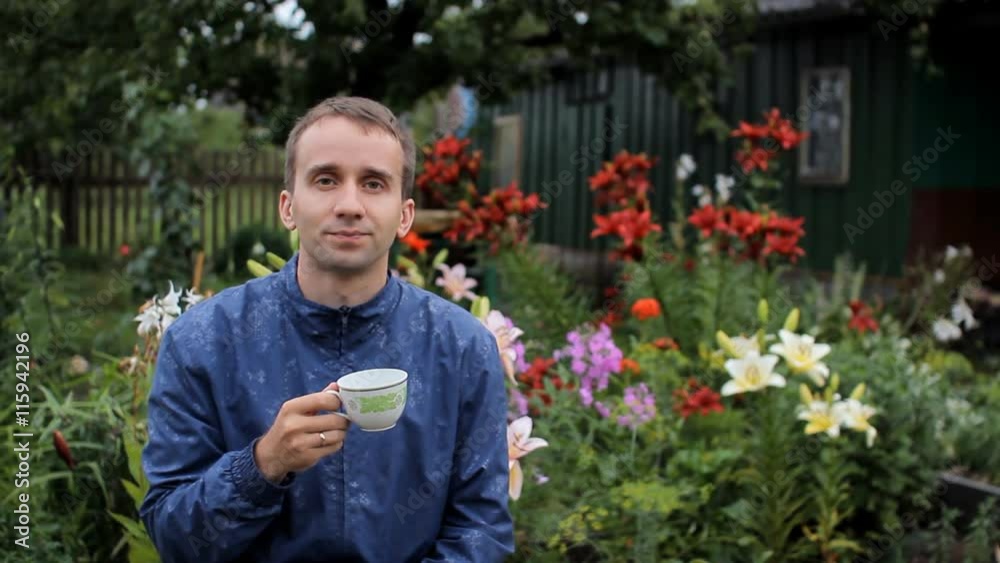Adult man drinks morning coffee in garden, summer day. Against the background of white, red, purple flowers Adult man drinks morning coffee in garden, summer day. Against the background of white, red, purple flowers