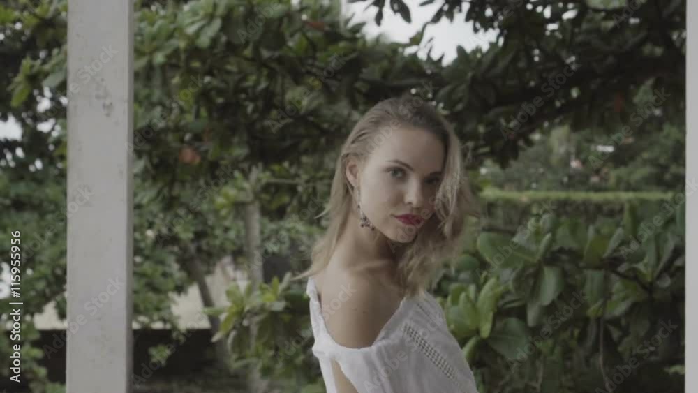 Back view of pretty naturally looking woman on summer day in boho style fashion wearing white shirt, bikini and earrings shaking hair on a terrace over green leaves background - video in slow motion. Back view of pretty naturally looking woman on summer day in boho style fashion wearing white shirt, bikini and earrings shaking hair on a terrace over green leaves background - video in slow motion.