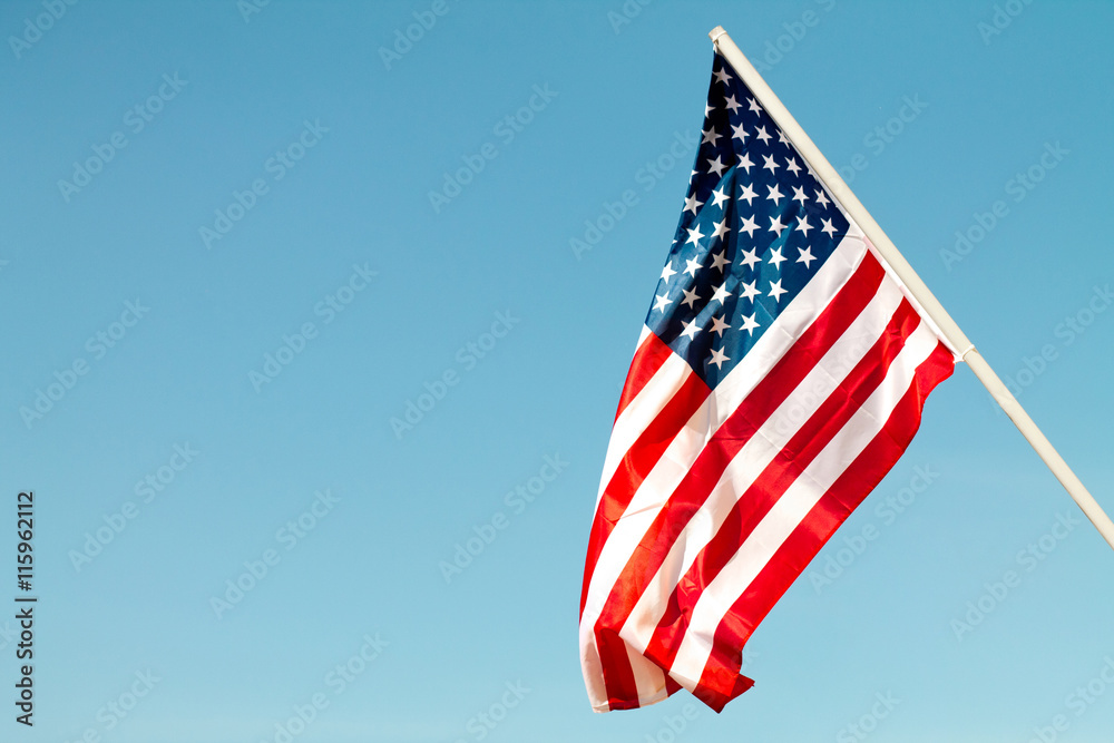 United States flag blows in the wind against a blue sky attached to the wall from the side. - obrazy, fototapety, plakaty 
